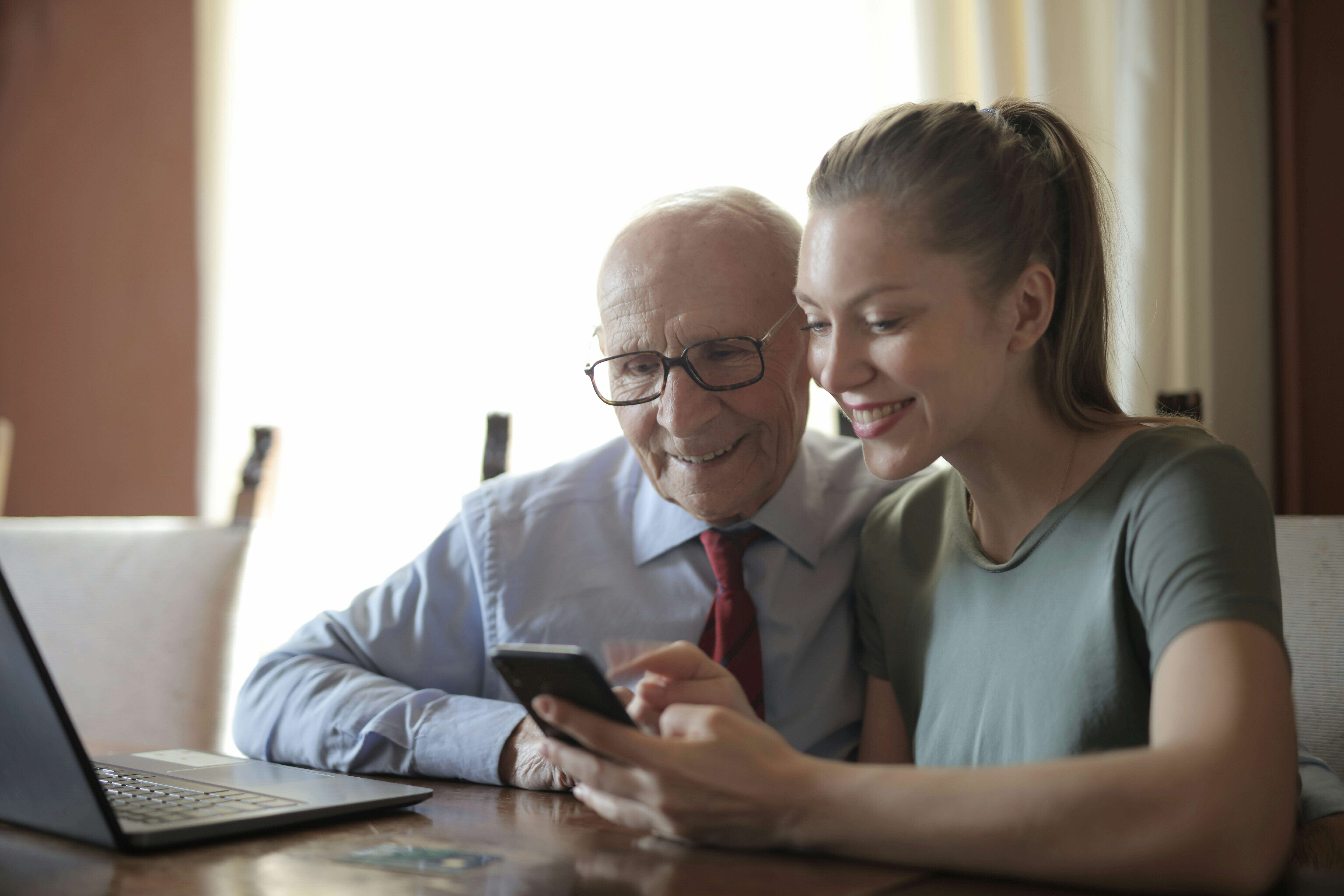 Grandfather and his granddaughter are sitting at the table in front of the laptop and looking at the smartphone that granddaughter is holding in her hands